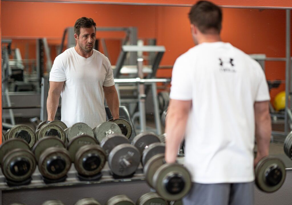 Man lifting dumbbells in a gym, facing a mirror. Orange wall, exercise equipment visible.