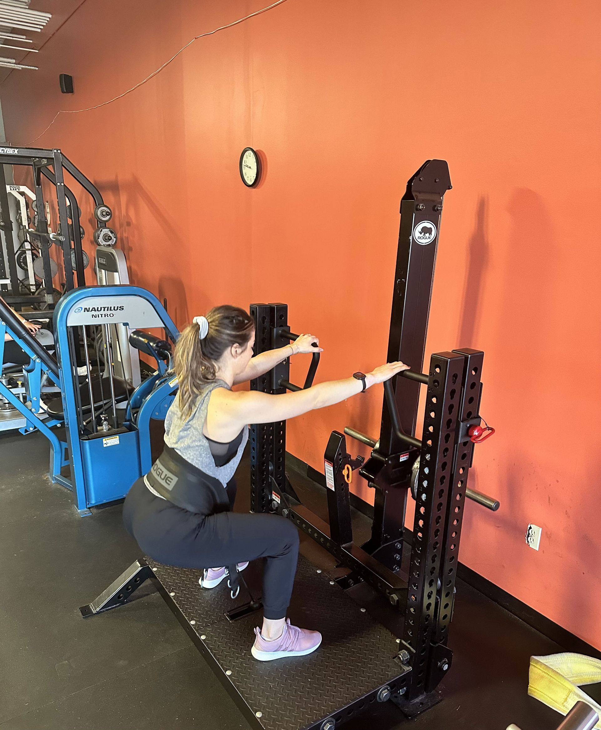 Woman squats on a black weight machine, handles in hand, in a gym with orange wall.