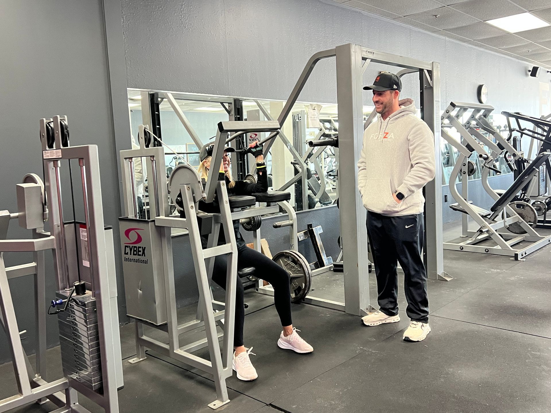 Woman using a seated gym machine, being coached by a man. They're in a gym with other equipment.