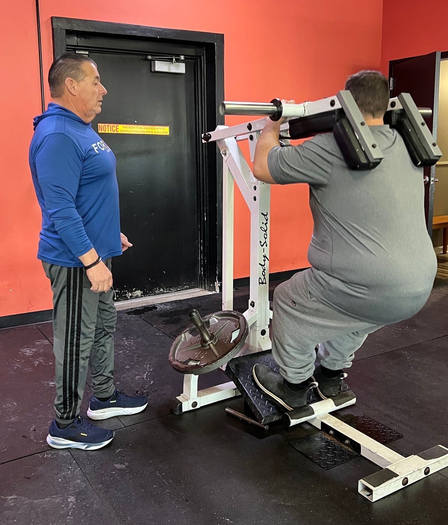 Man using a squat machine while another man watches in a gym with red walls.