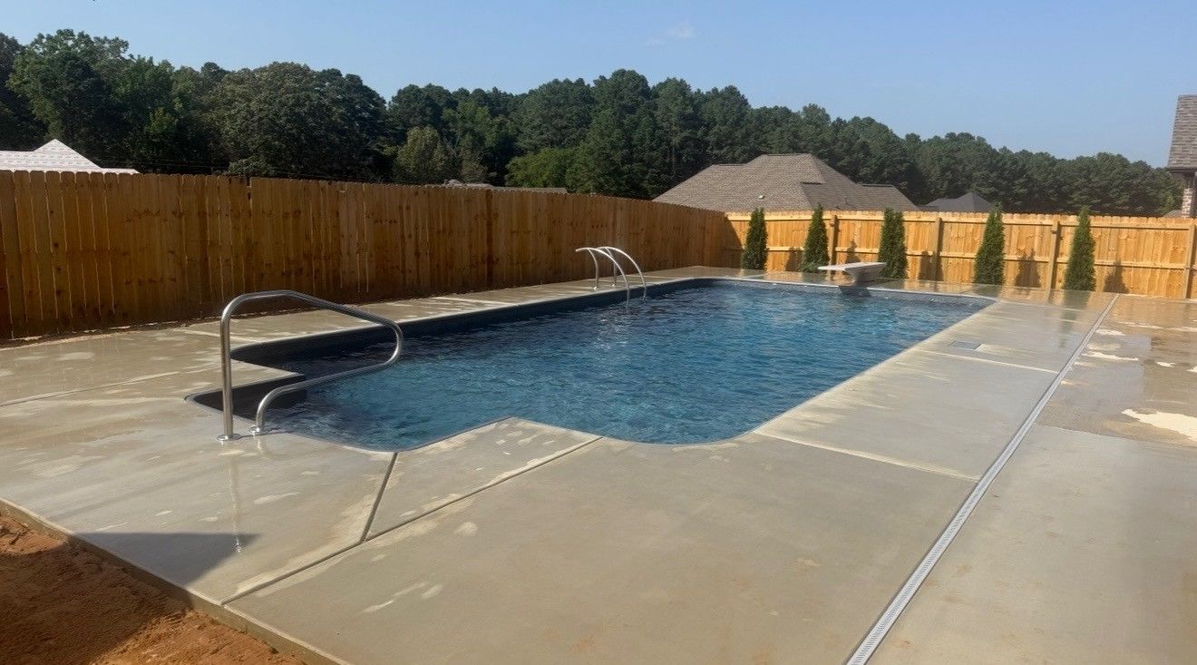 Outdoor backyard swimming pool with blue water, concrete deck, and wooden privacy fence under clear sky