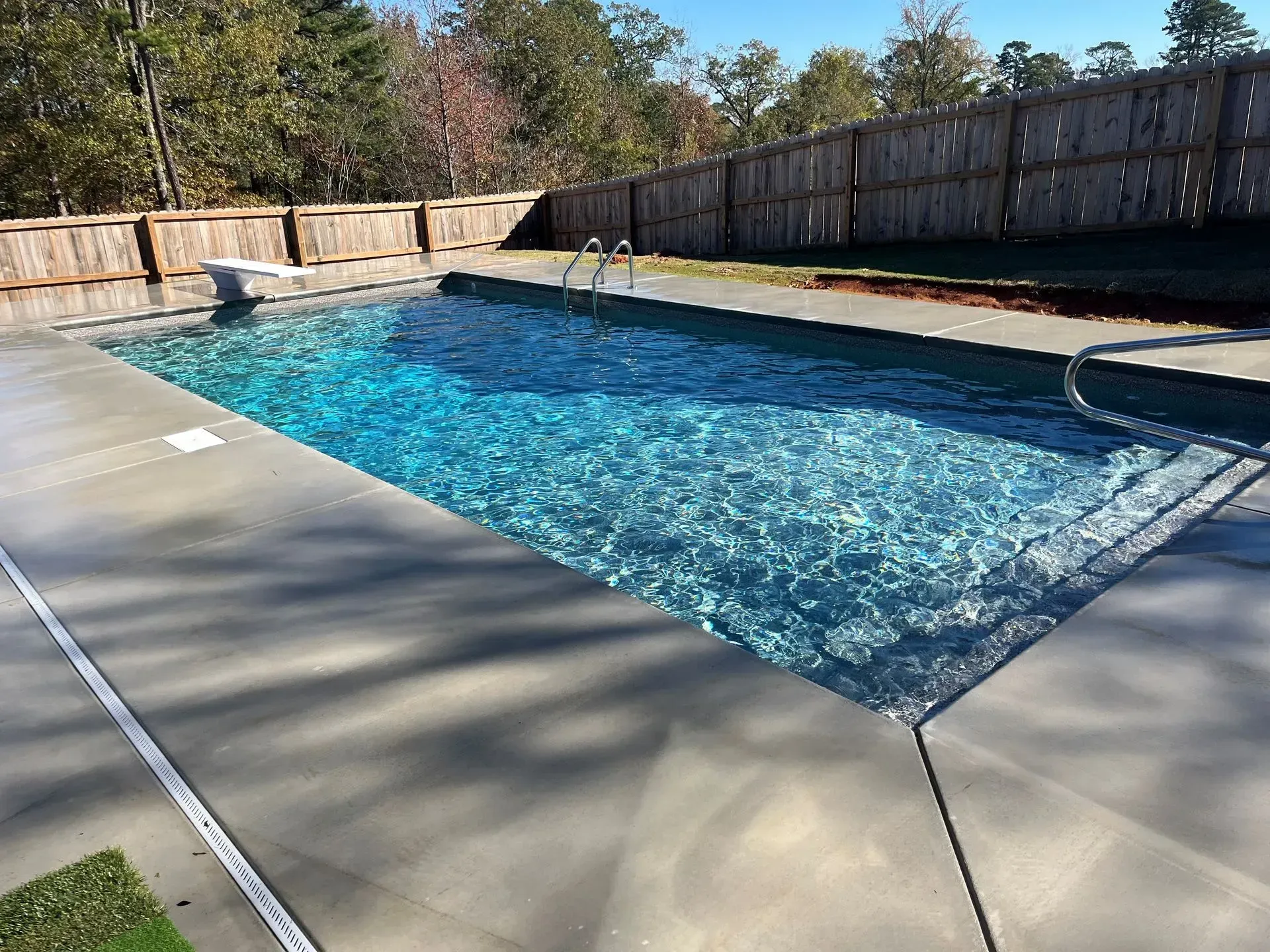 Backyard swimming pool with clear blue water, concrete deck, and wooden fence in sunlight