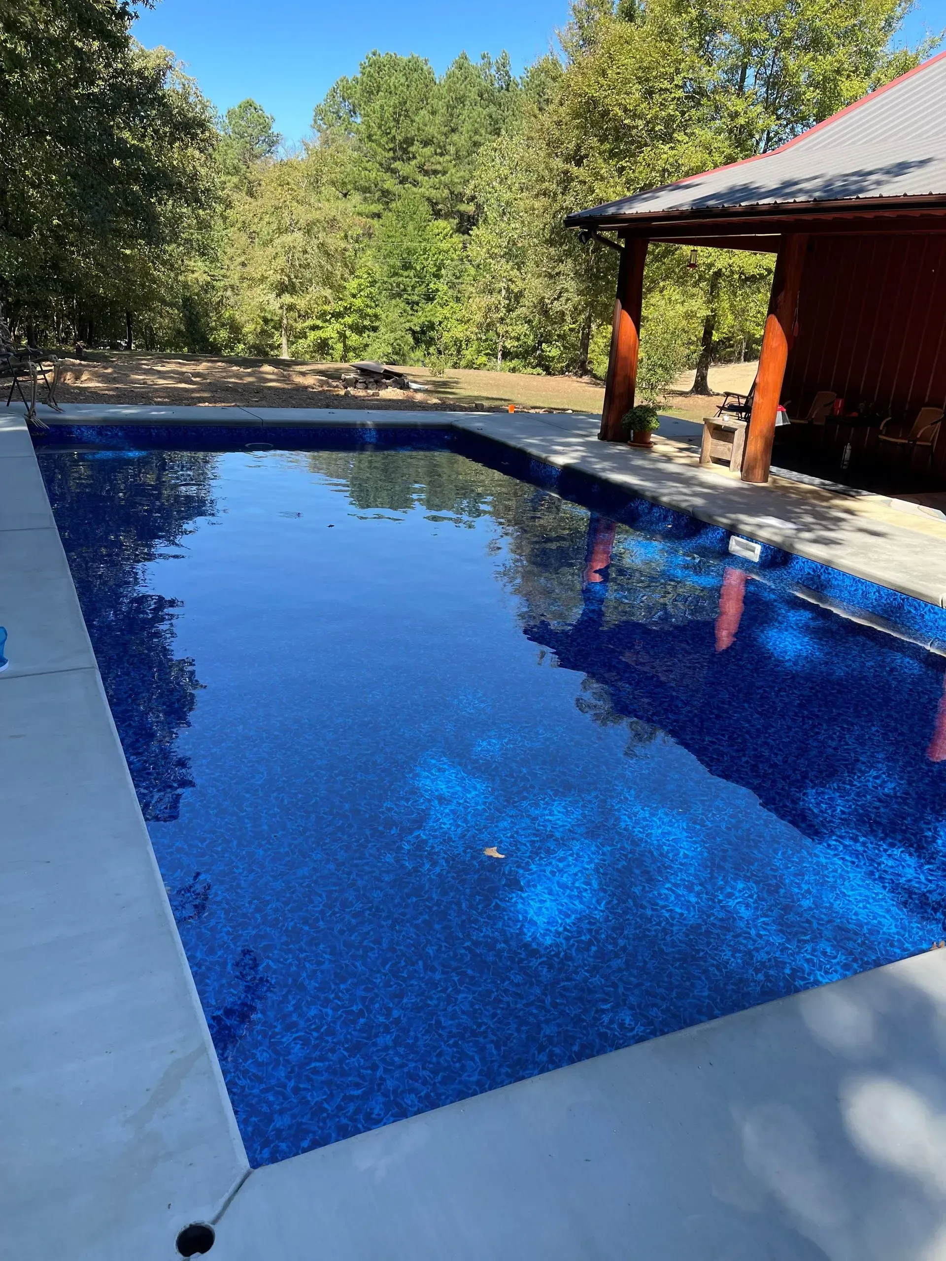 Blue tiled swimming pool beside a covered patio, with trees and bright sky reflected in the water