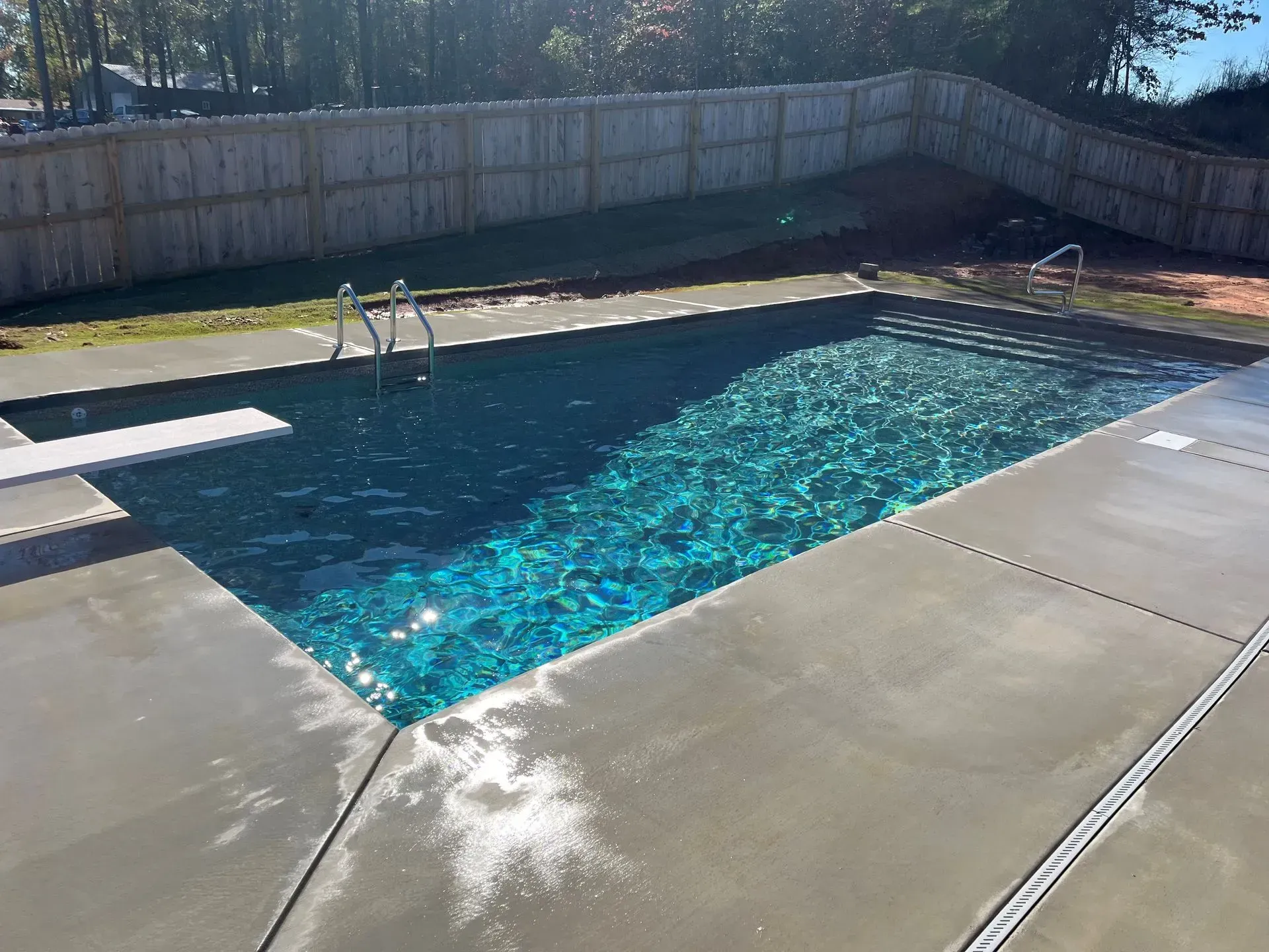 Backyard swimming pool with blue water, concrete deck, and wooden fence in sunlight