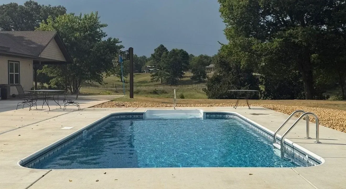 Rectangular outdoor swimming pool with blue water beside a house and trees under a clear sky