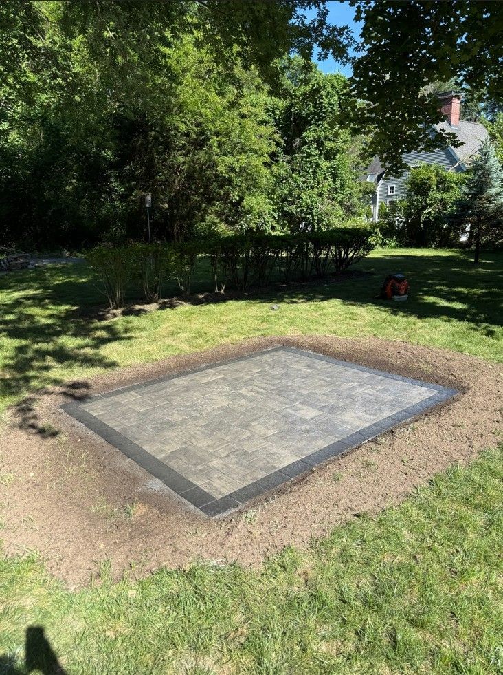 Small concrete slab bordered by brick in a grassy backyard with trees and a house in the background