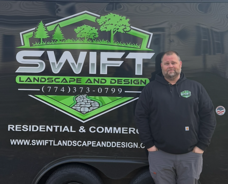 Man standing by a yellow stump grinder on a driveway, wood chips scattered around.