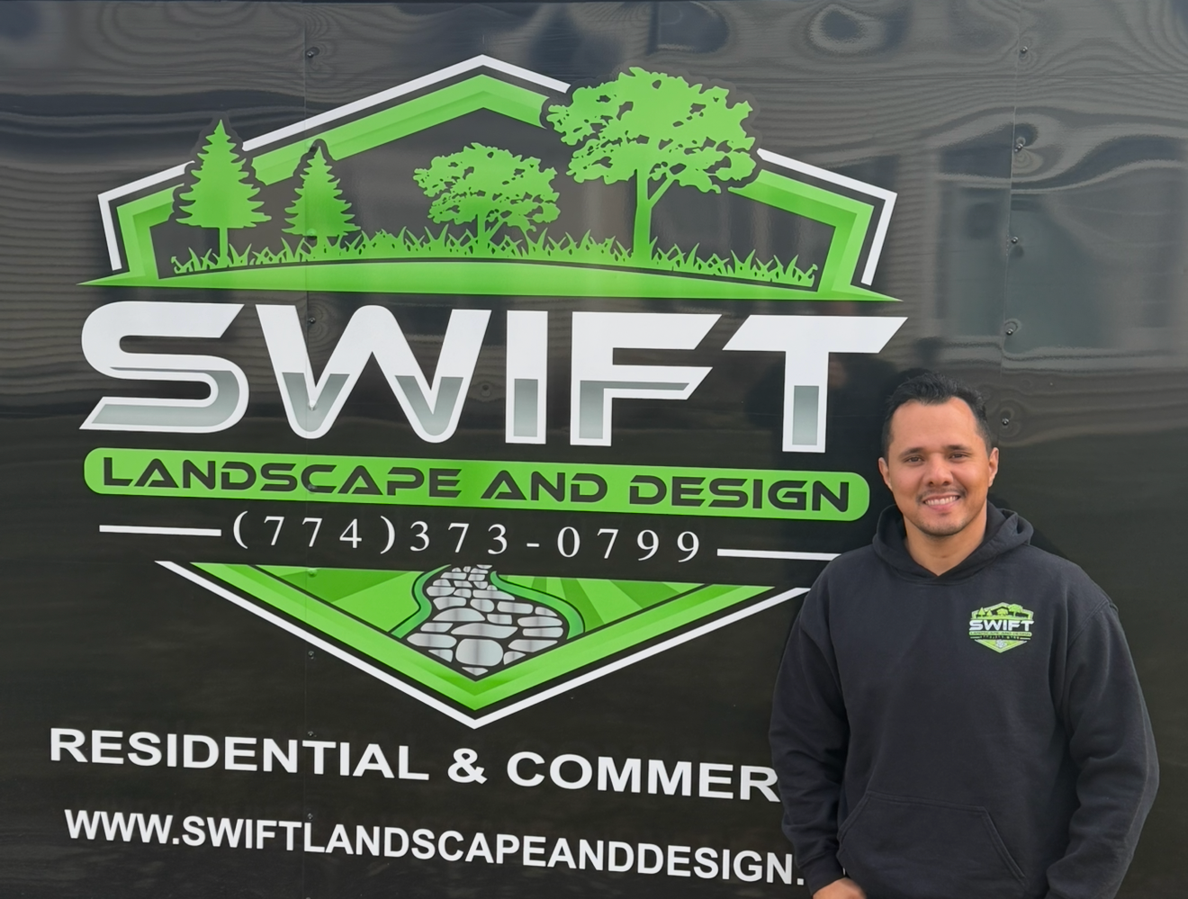 Man in black hoodie stands outside a building. Hands in pockets. Sign for 