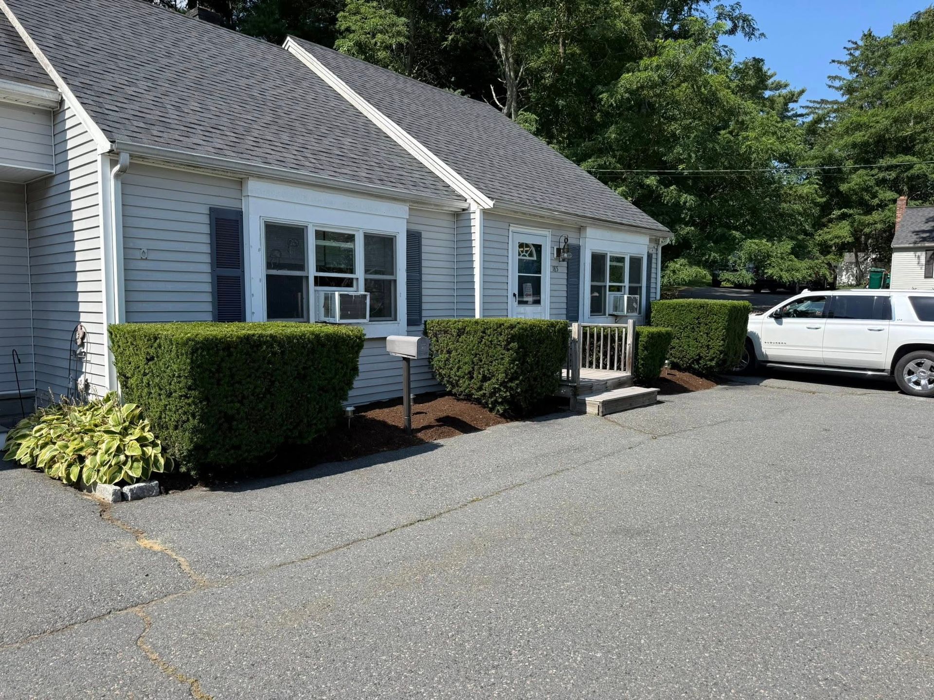 Small, light-colored building with dark roof, windows, and manicured shrubs. Asphalt driveway, white SUV parked nearby.
