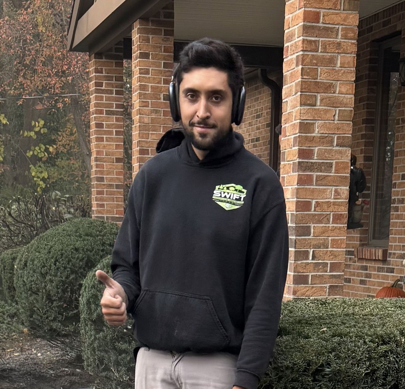 Man wearing headphones, thumb up, standing in front of a brick building and bushes, smiling.