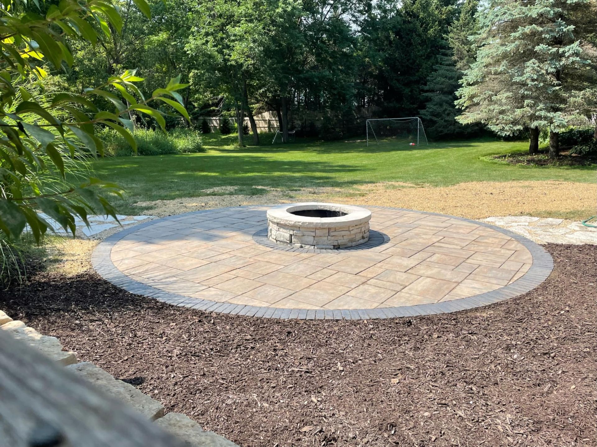 Circular brick patio with fire pit, surrounded by mulch and grass, trees in background.