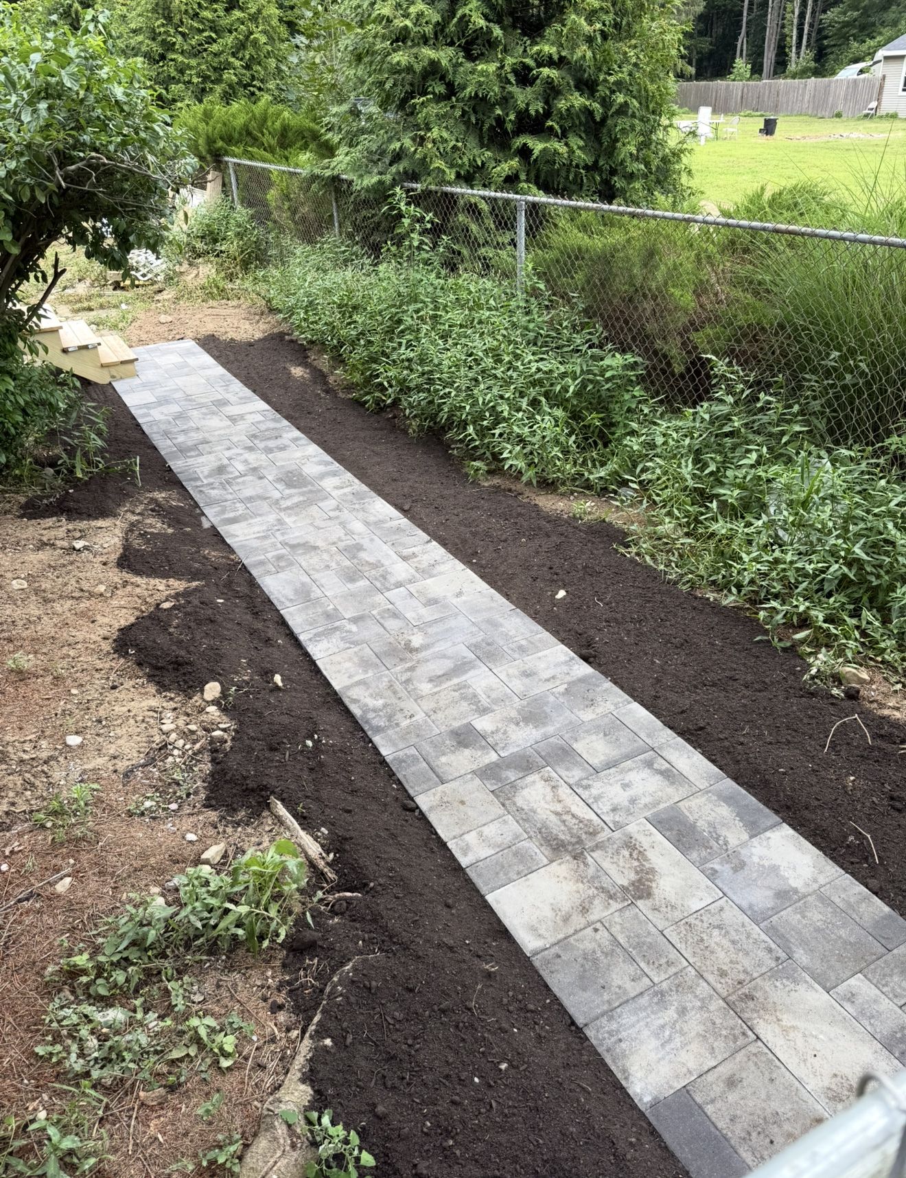 Paver walkway in a yard with dark soil on both sides, surrounded by greenery and a chain-link fence.