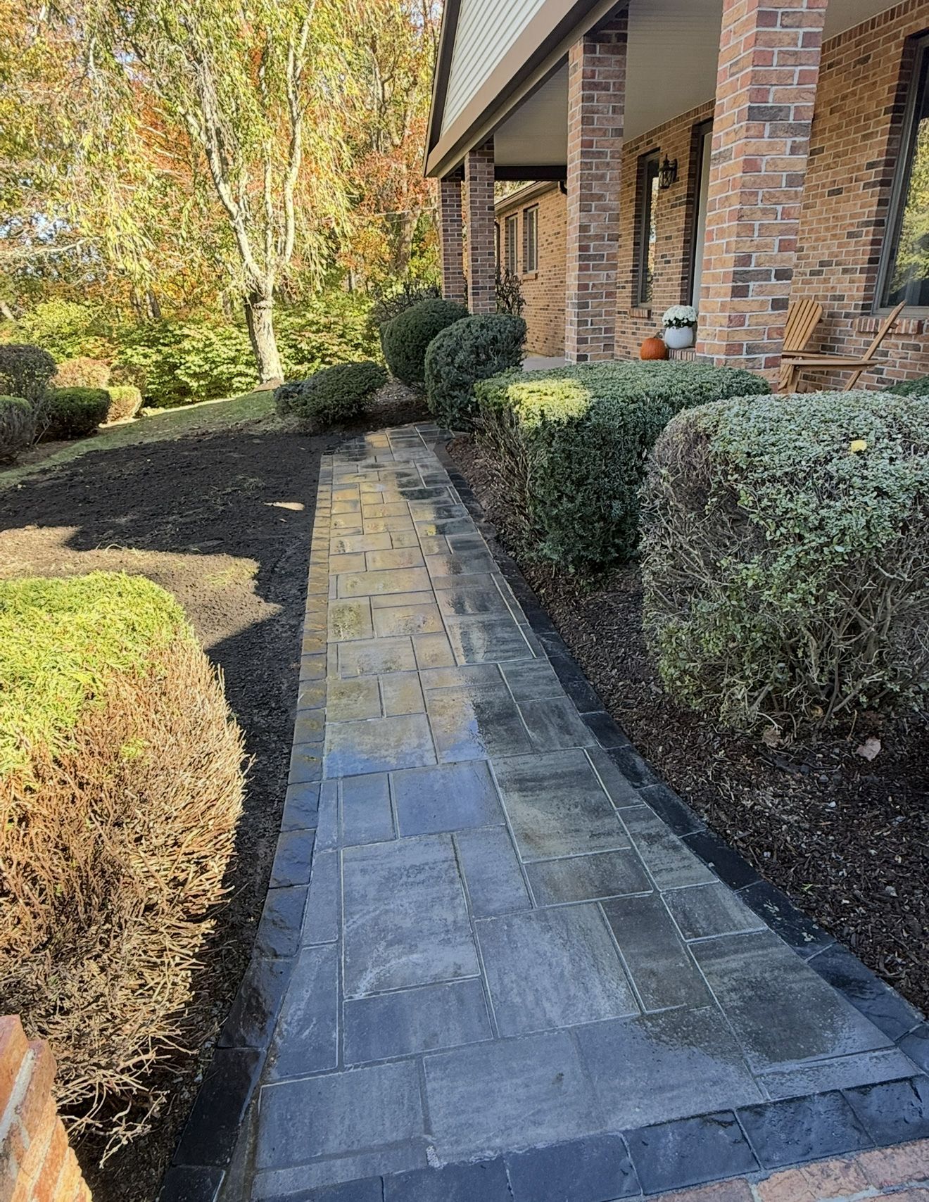 Brick pathway leading to a house with hedges and a porch, set outdoors with trees.