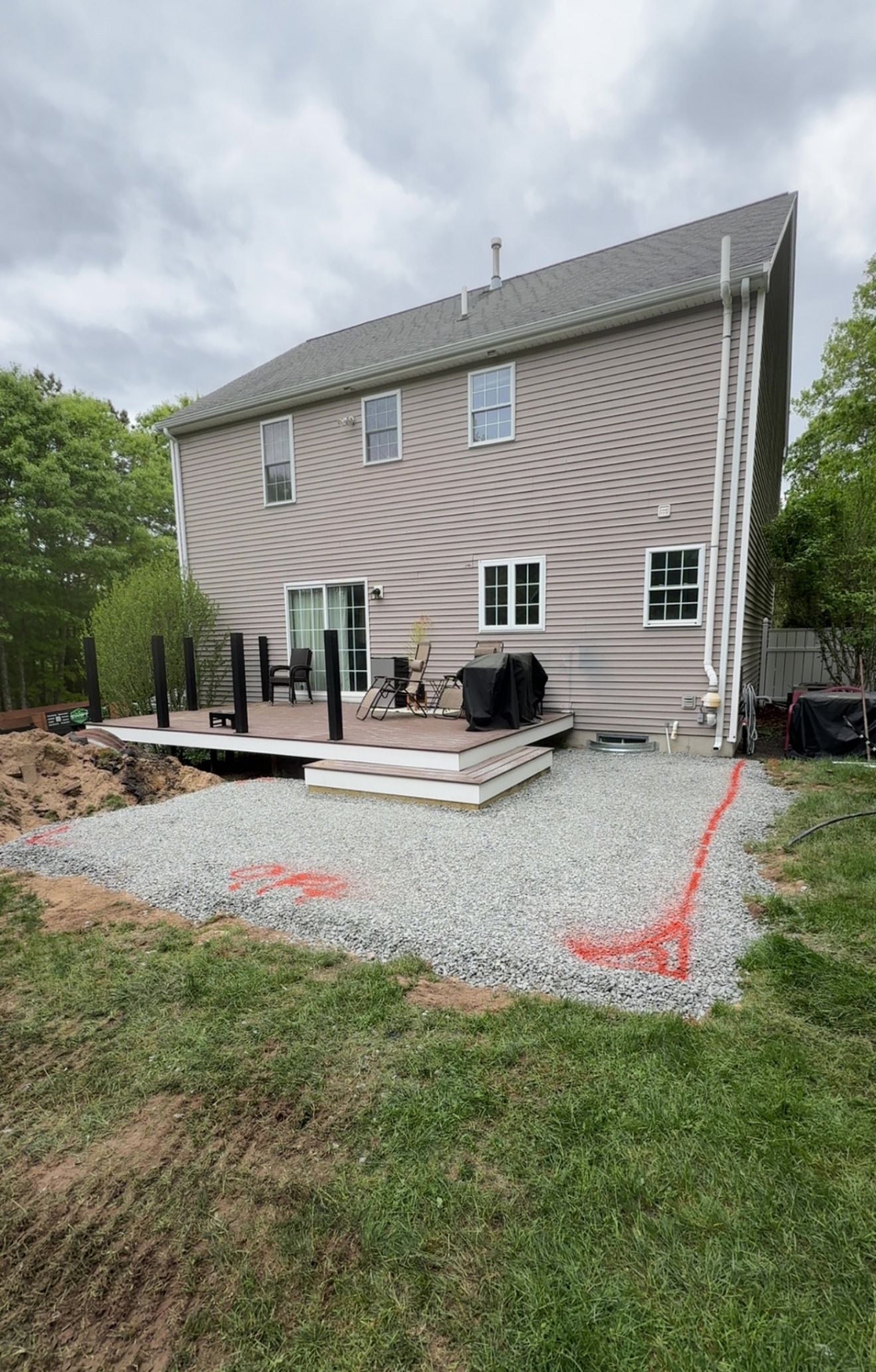 Back of house with a deck, gray siding, and gravel patio in progress, marked with orange paint.