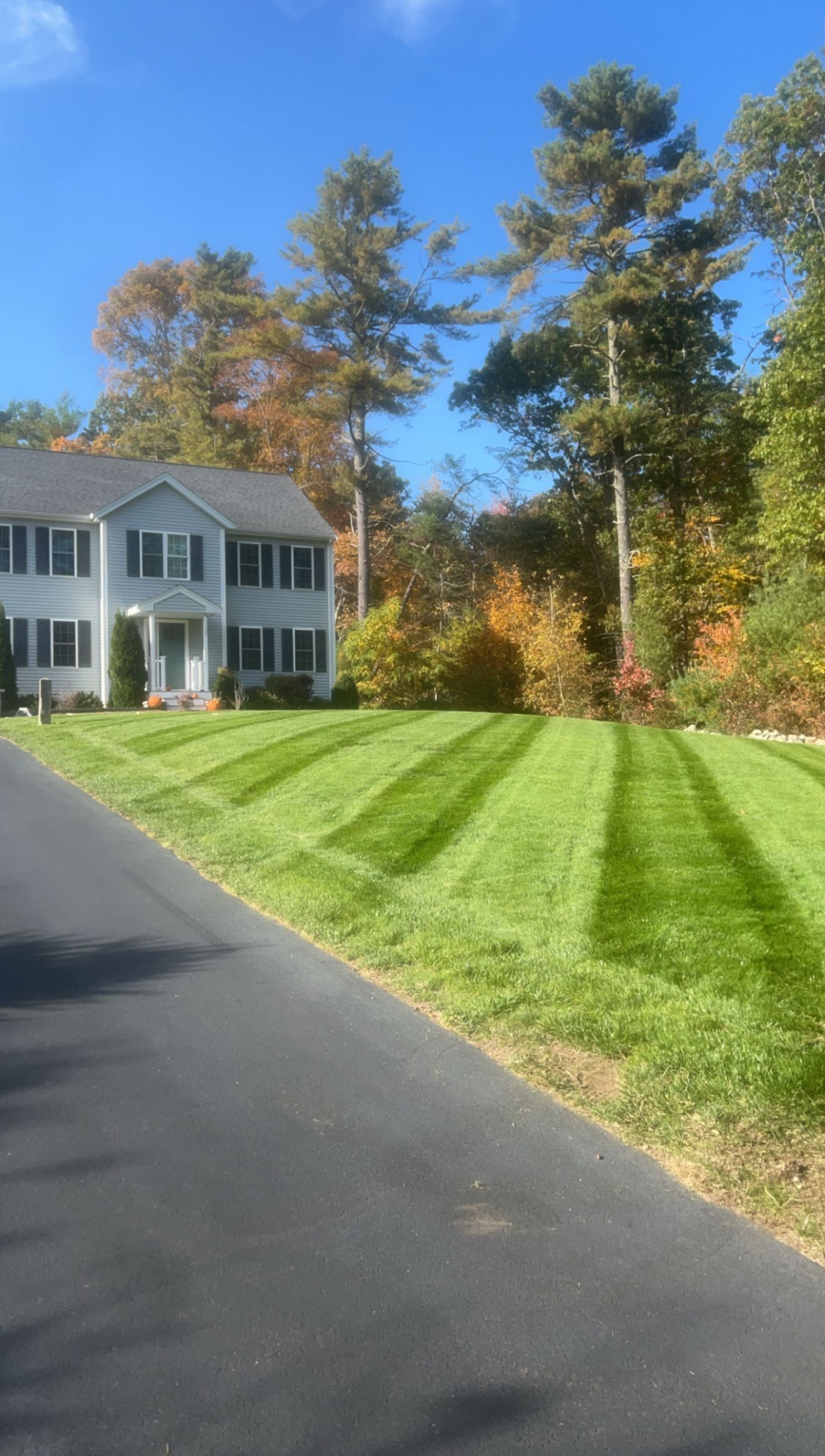 Two-story house with green lawn striped by a mower, trees with fall foliage in background, and asphalt driveway.