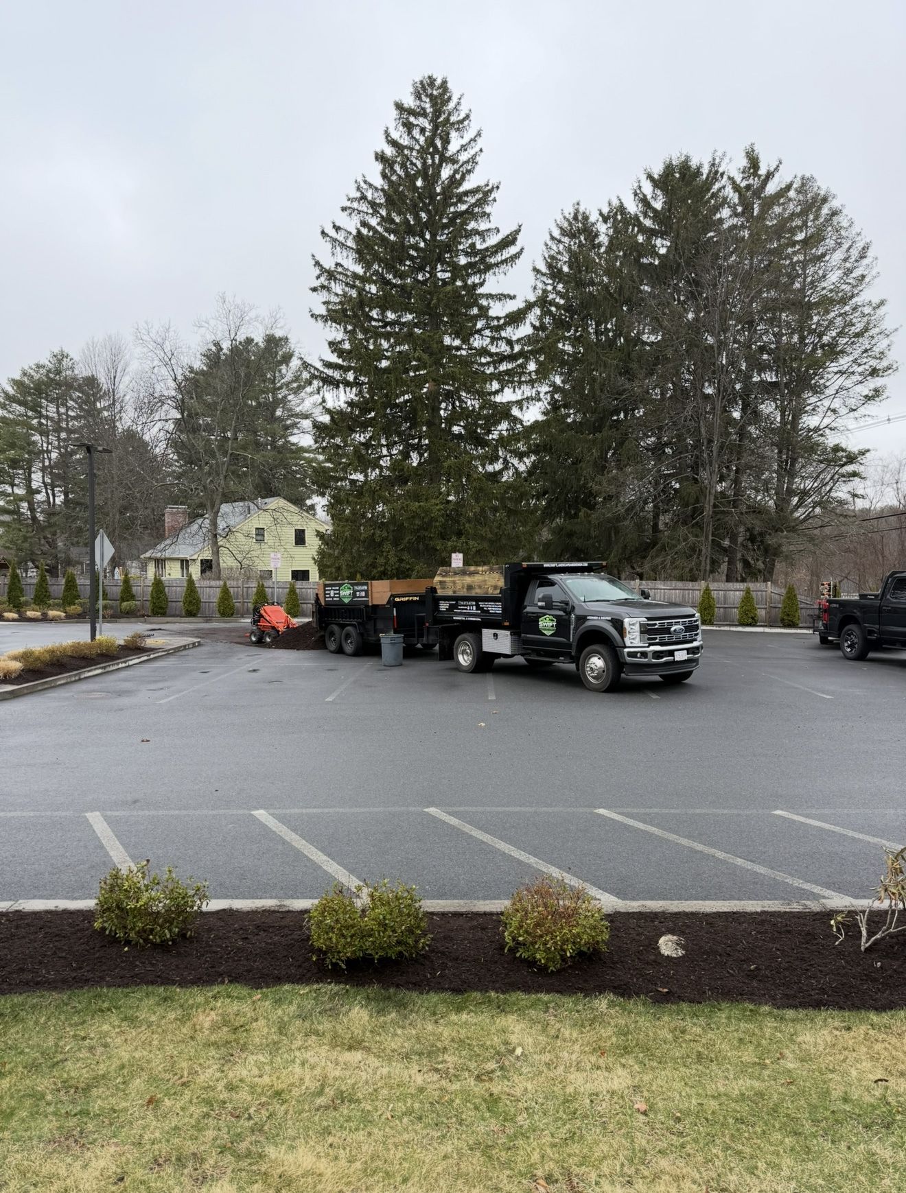 A work truck and trailer loaded with landscaping materials parked in an asphalt lot. Shrubs line the foreground.