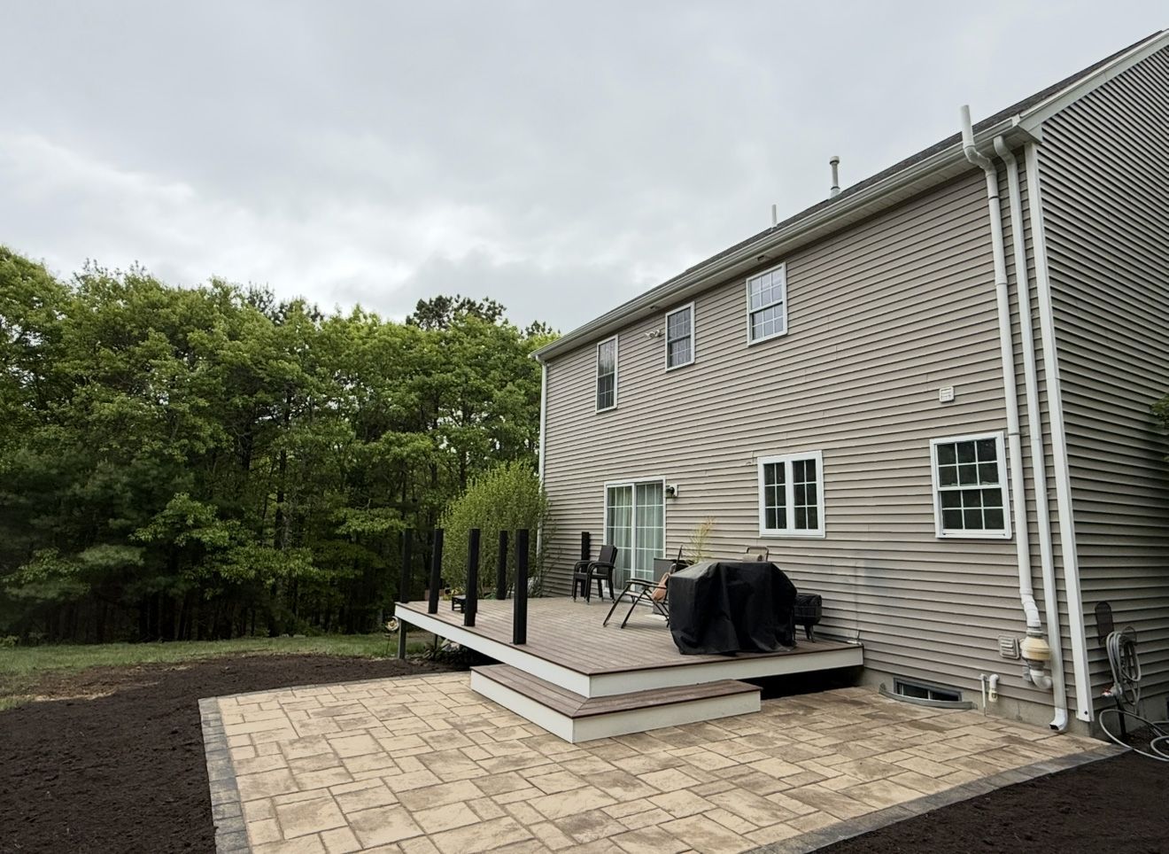 Back of a two-story beige house with a deck and brick patio. Overcast sky with trees in the background.