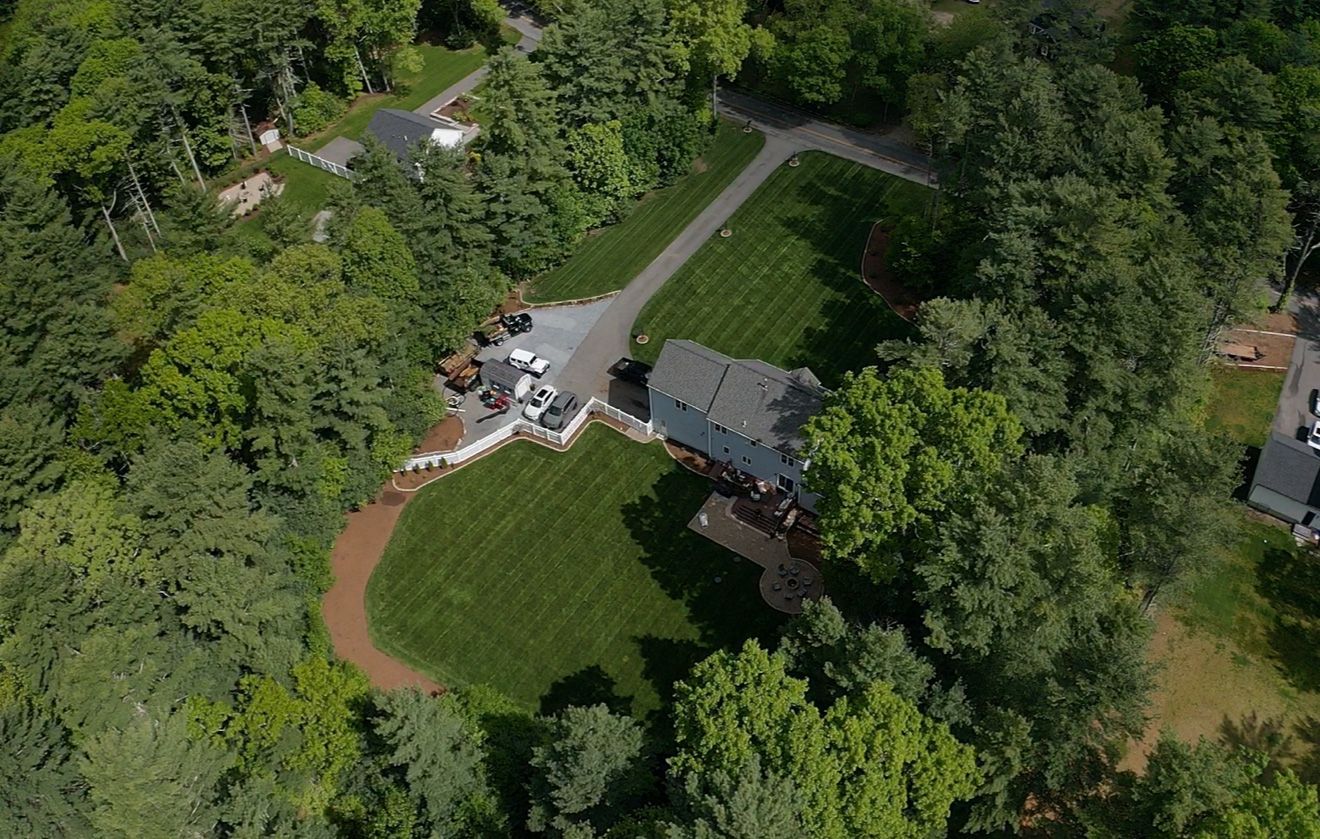 Aerial view of a gray house with a green lawn, surrounded by trees and a long driveway.