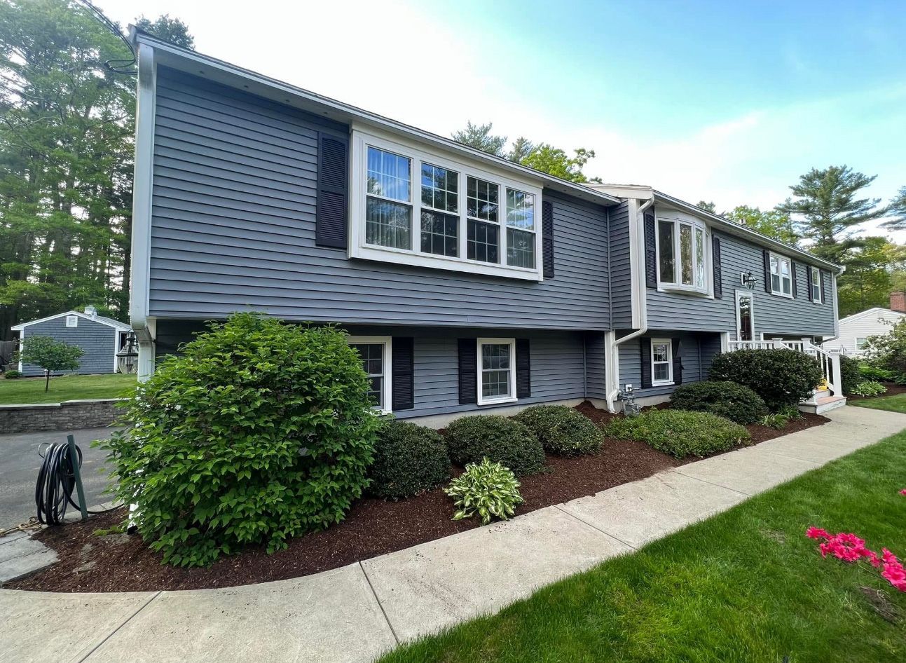 Two-story house with dark blue siding, bay windows, and a well-manicured yard with bushes and a sidewalk.