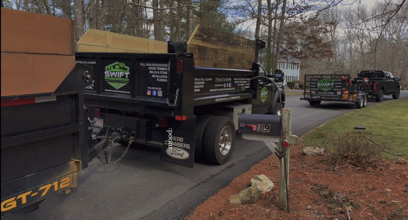 Black dump trucks and trailer parked on a road, loaded with materials; a residential area.