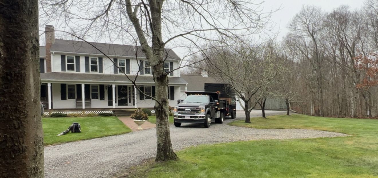 Truck and trailer parked on a gravel driveway in front of a large white house. Overcast sky.