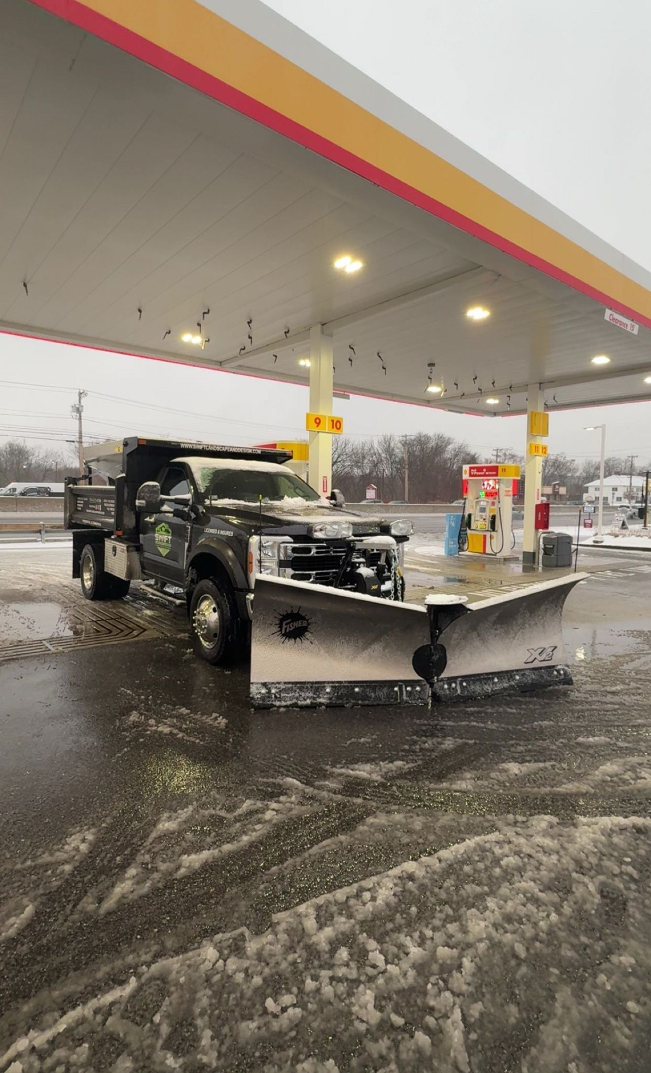 Snowplow truck at a Shell gas station. Snow covers the ground and the truck's blade. The sky is overcast.