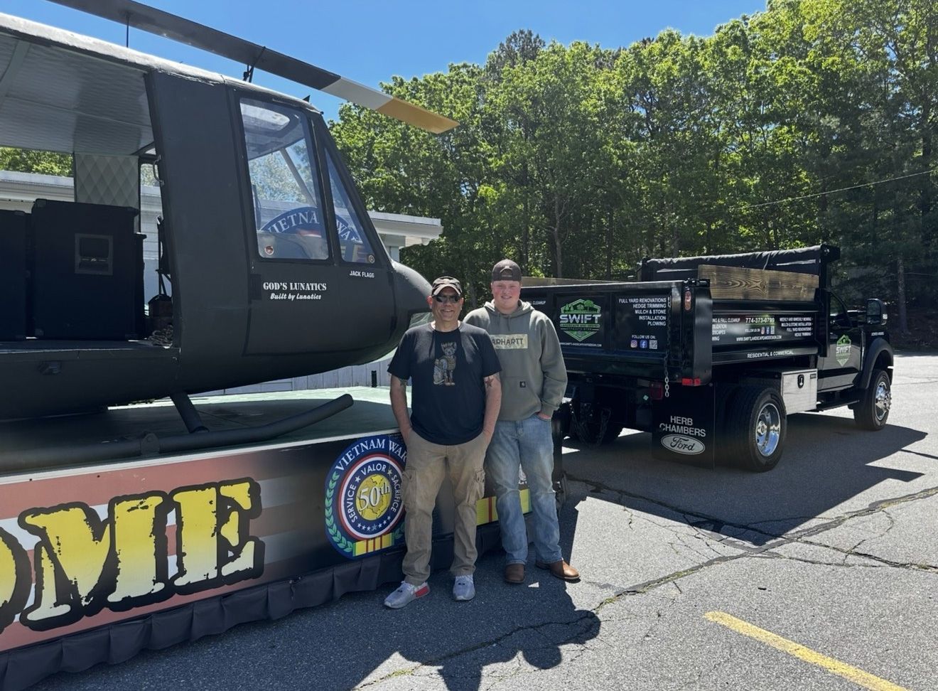 Two men stand by a military helicopter display on a parade float and a dump truck.
