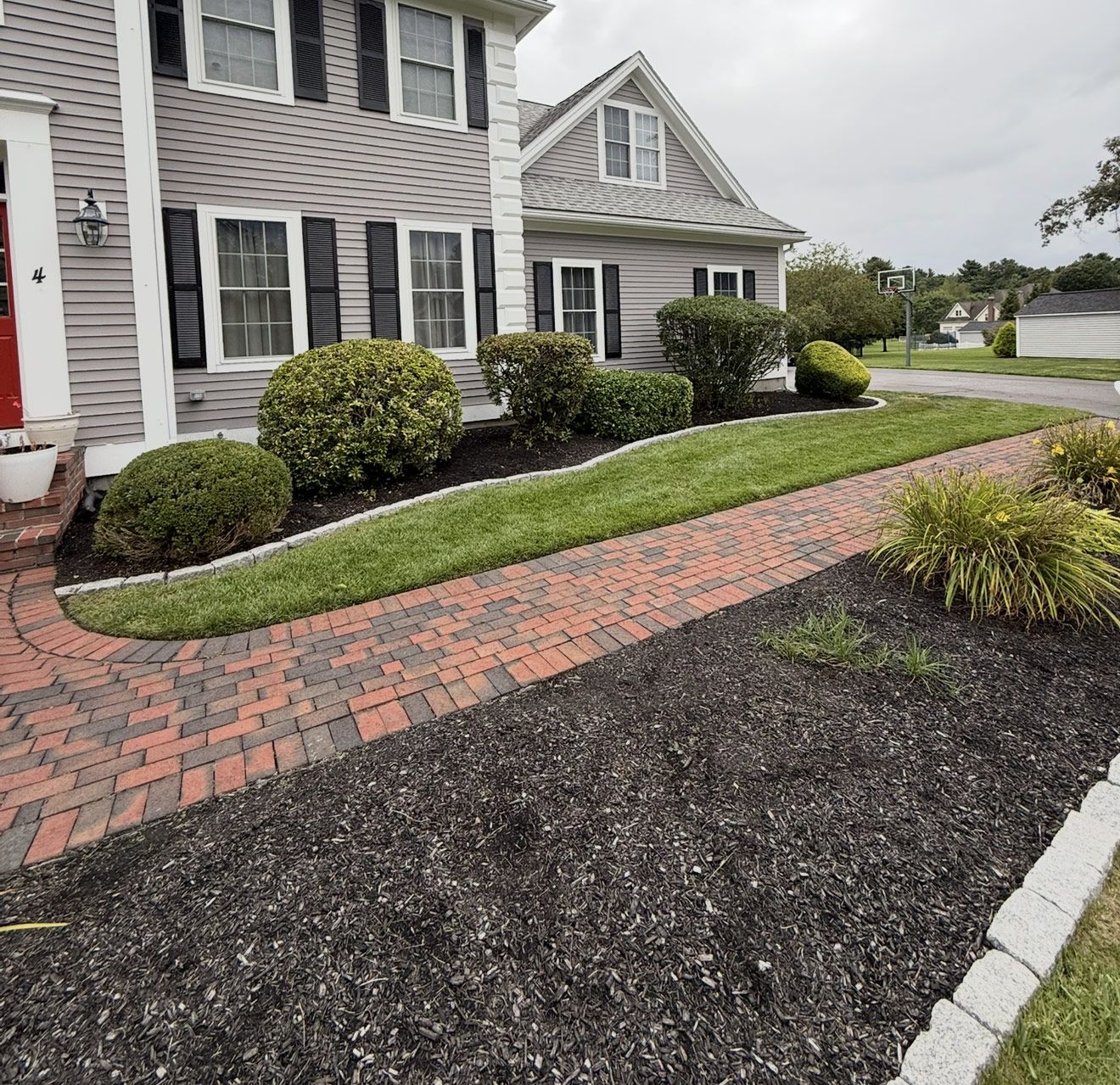 Brick walkway leads to a house with gray siding, black shutters, and well-maintained landscaping.