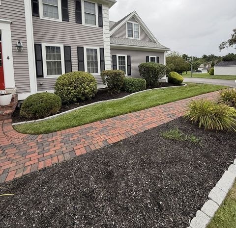 Brick walkway leads to a house with gray siding, black shutters, and well-maintained landscaping.