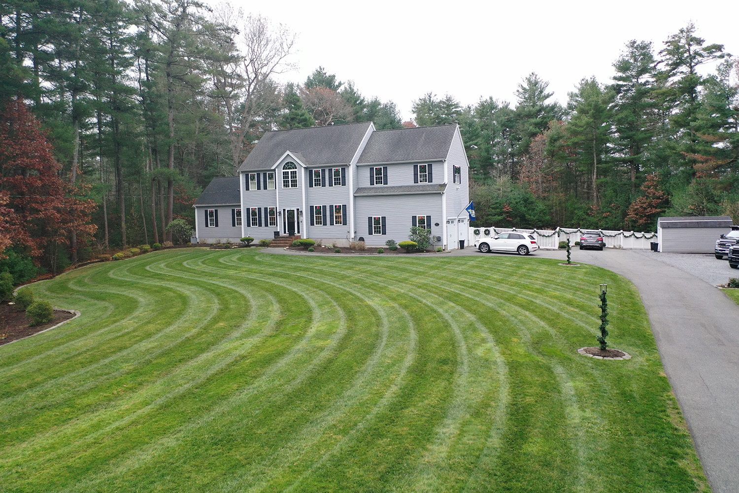 Two-story gray house with green lawn featuring curved mowing patterns. Cars in driveway and surrounding trees.
