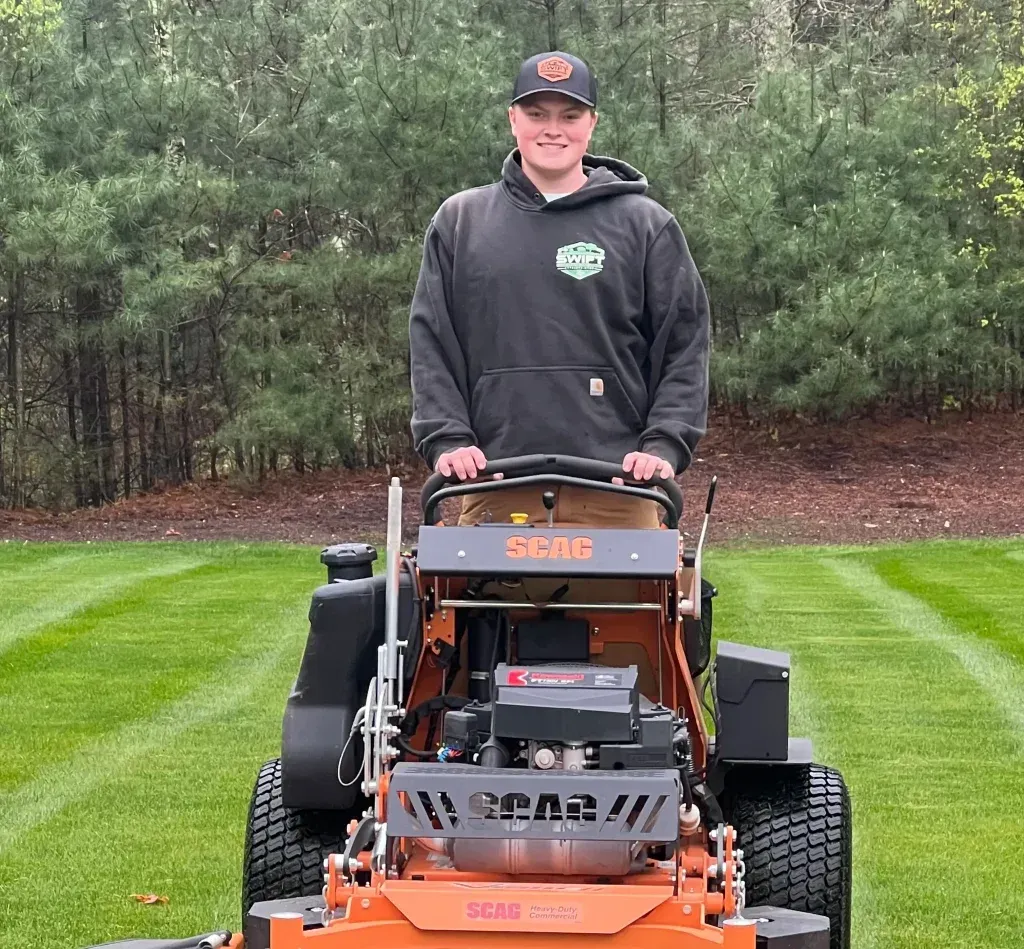 Person operating orange lawn mower on a green lawn, wearing a black hoodie and baseball cap.