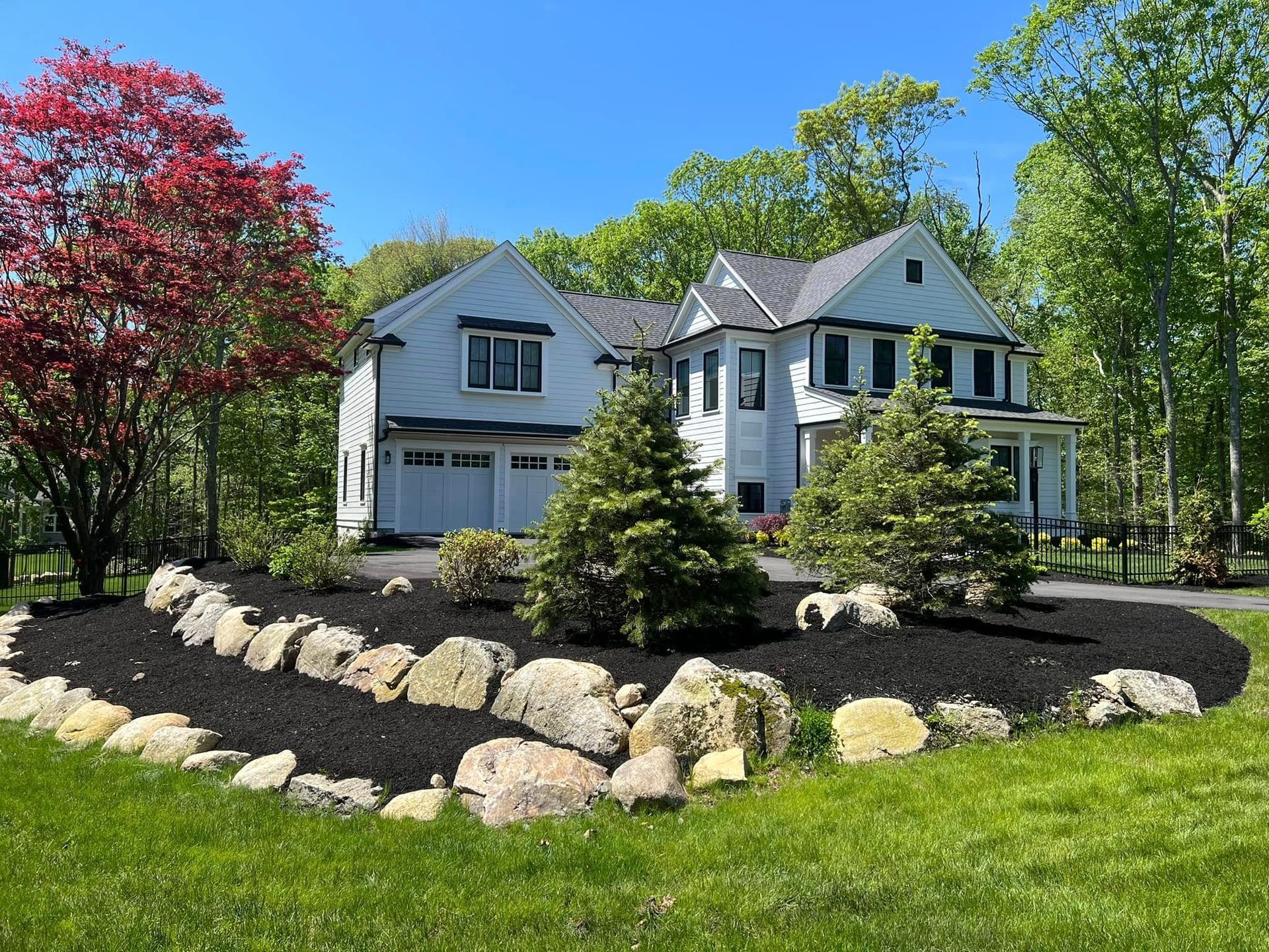 White house with black mulch landscaping, large rocks, and trees. Sunny day.