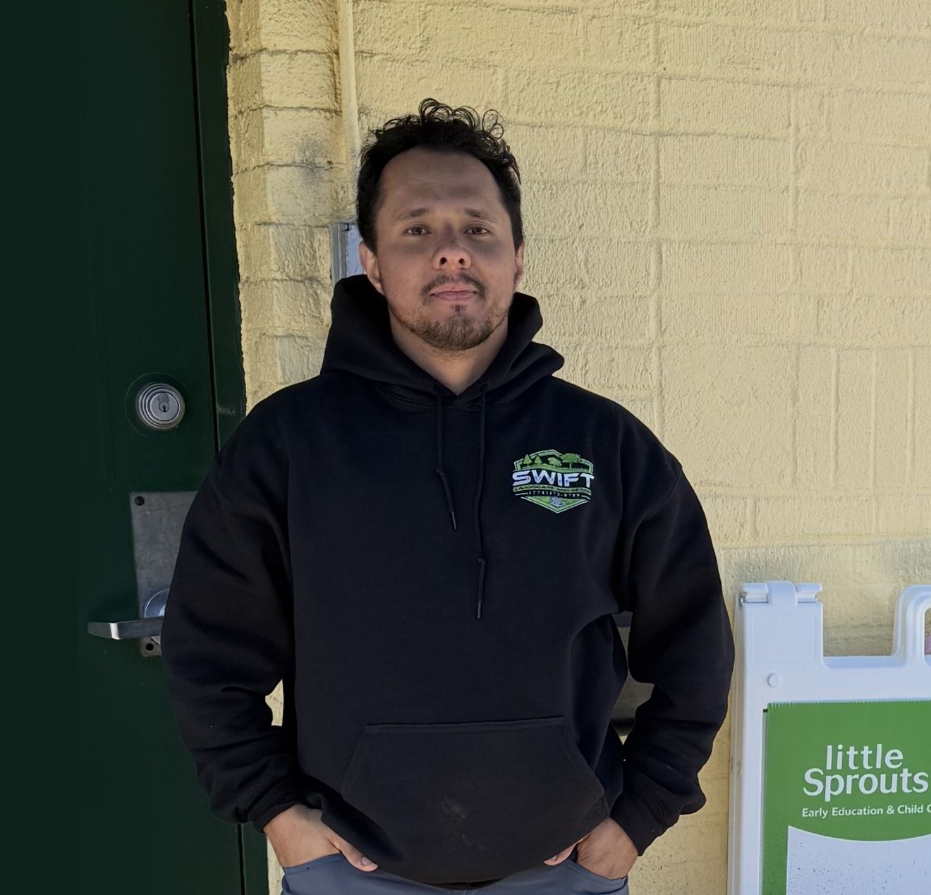 Man in black hoodie stands outside a building. Hands in pockets. Sign for 
