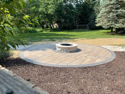 Circular stone patio with fire pit, surrounded by mulch and lawn. Trees in background.