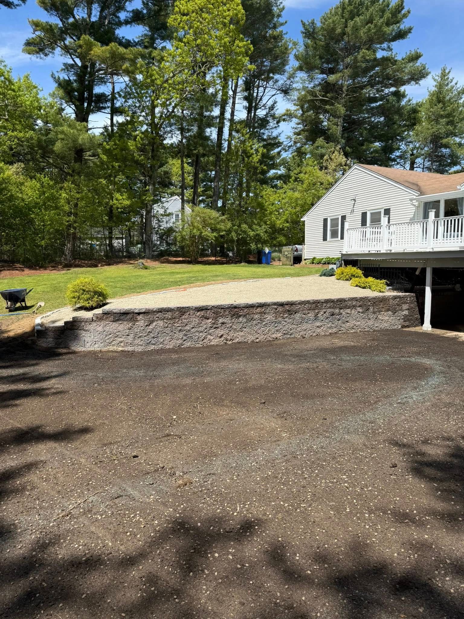 Yard with a low stone retaining wall, newly seeded area, and a house with a deck.