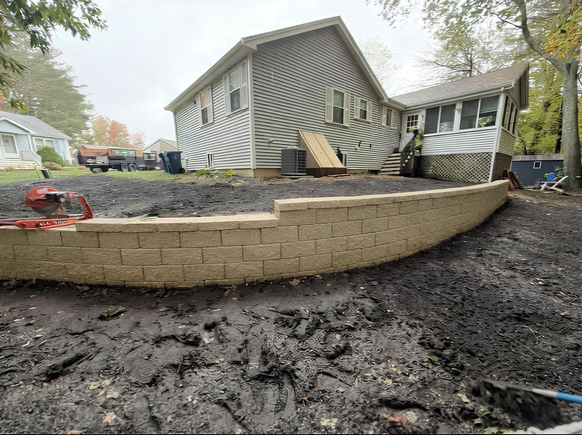 A backyard with a newly built curved retaining wall and a house in the background. Muddy ground.