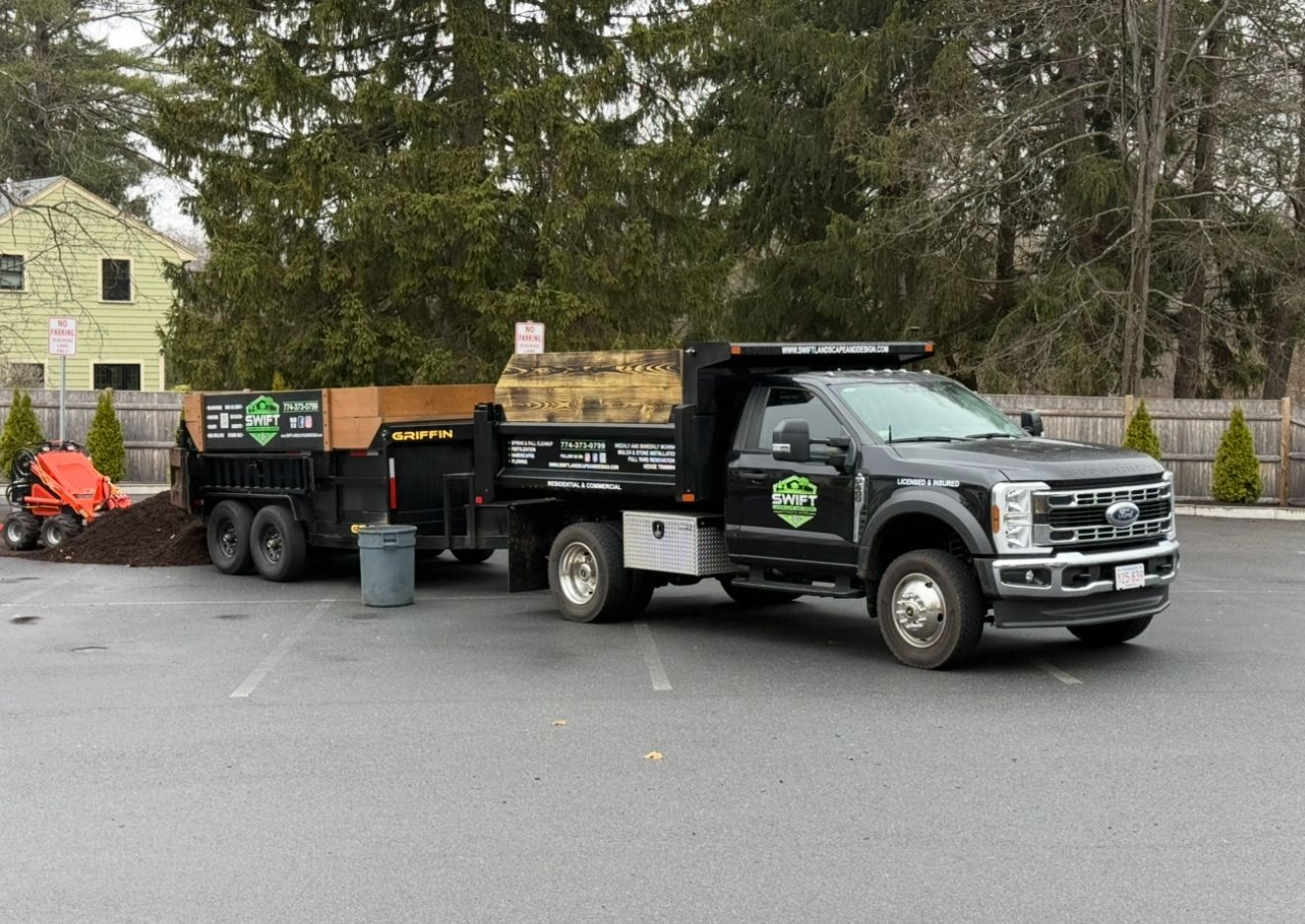 Black dump truck with trailer, parked in a lot, loaded with material; small tractor nearby.