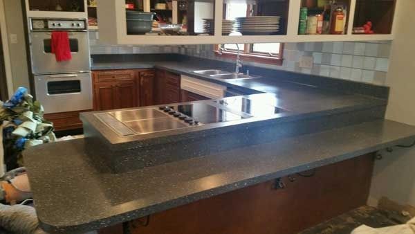 Kitchen with large black countertop island, stainless sink, wooden cabinets, and tiled backsplash