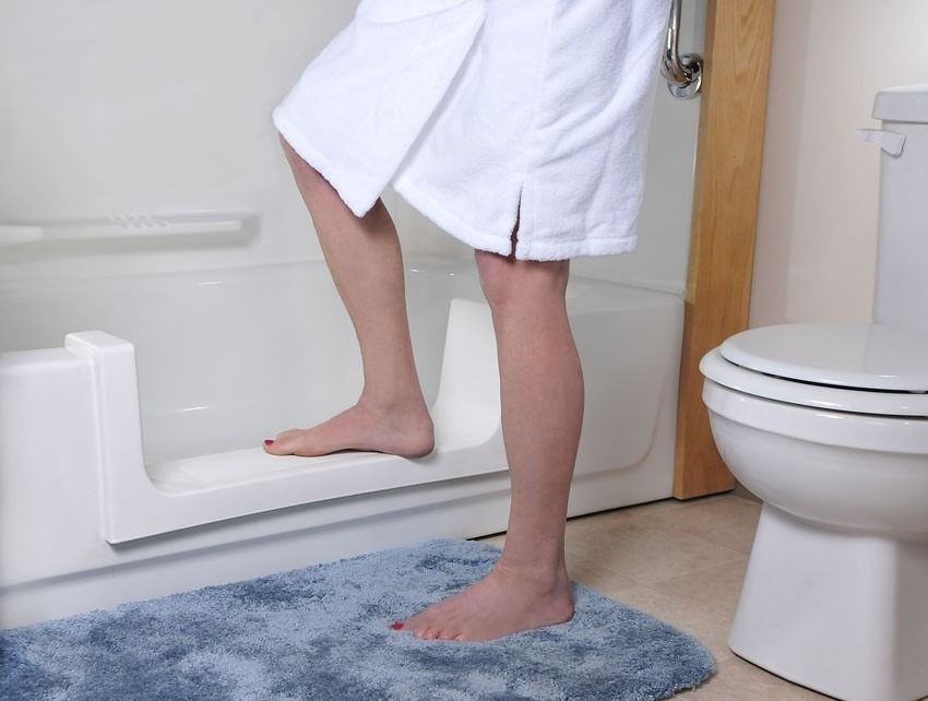 Person stepping into a white bathtub using a built-in low-step, beside a toilet and blue bath mat