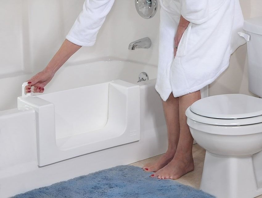 Person lowering into a white walk-in bathtub beside a toilet in a bathroom