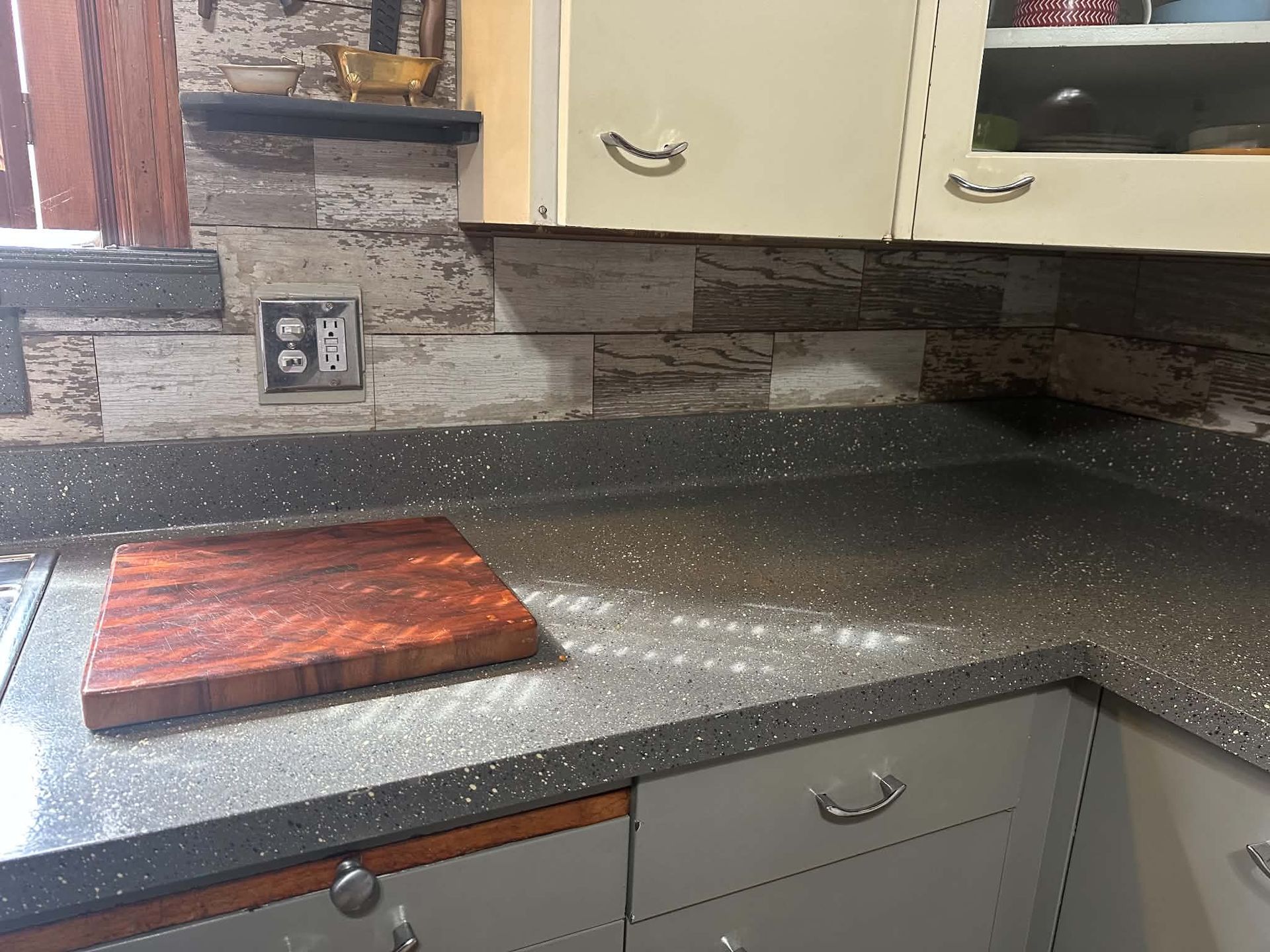 Kitchen countertop with wood cutting board, stone backsplash, and light gray cabinets