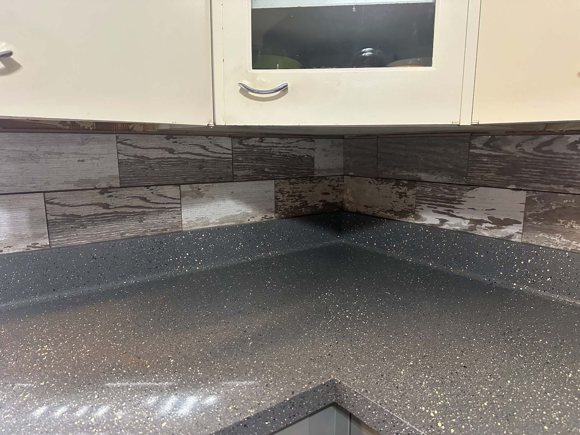 Speckled dark kitchen countertop with gray tile backsplash and white cabinets above.