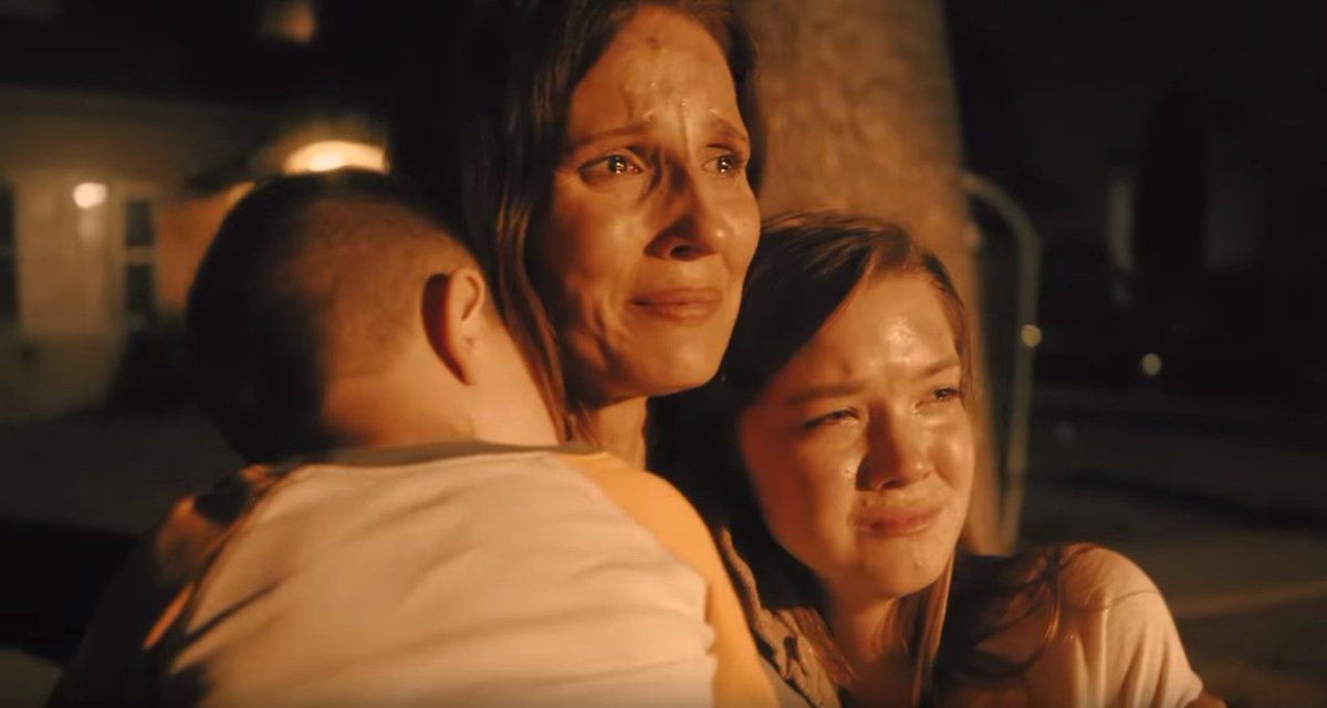 Woman hugs child and teen, all with tearful expressions outdoors at night.