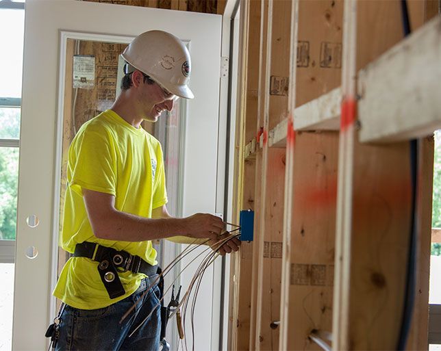 Construction worker in a yellow shirt and hard hat wiring electrical cables in a wooden wall.