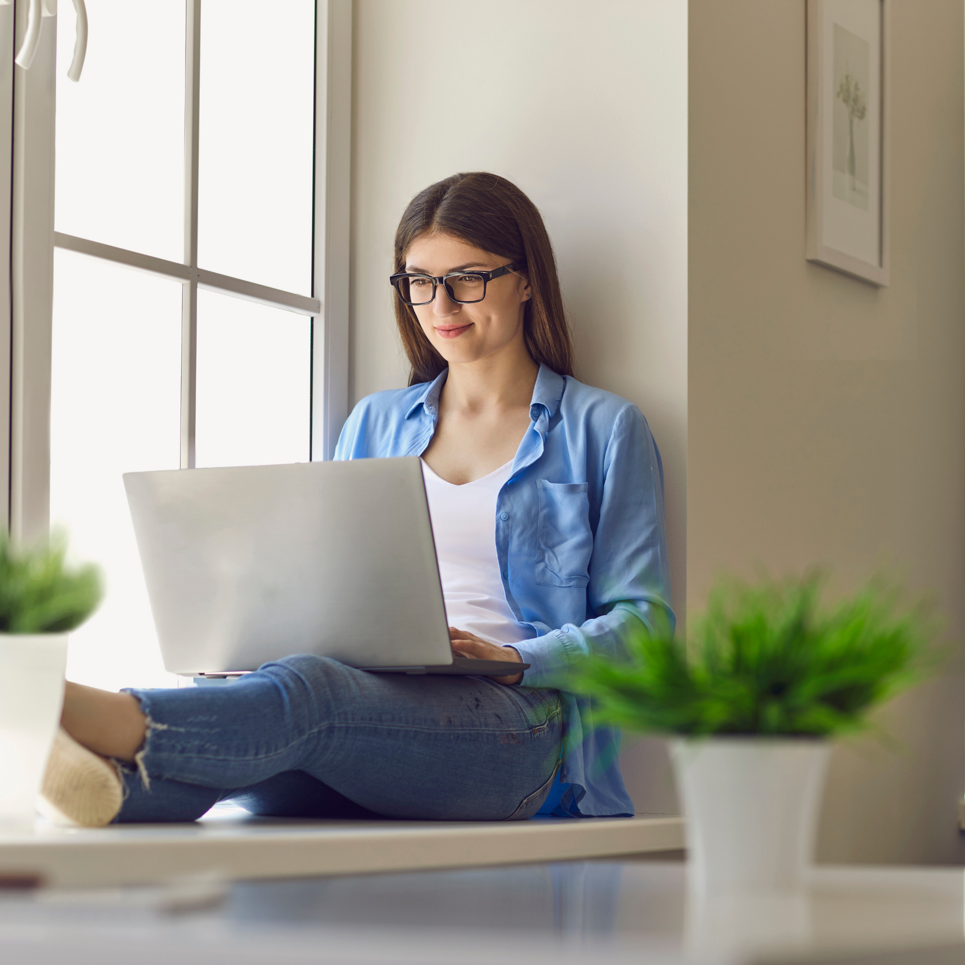 Woman in glasses using laptop, sitting in a window seat, wearing a blue shirt and jeans.