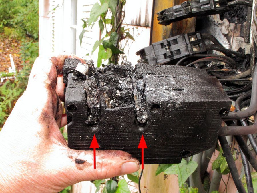 Hand holding a charred electrical component with melted wires. Red arrows point to mounting holes.