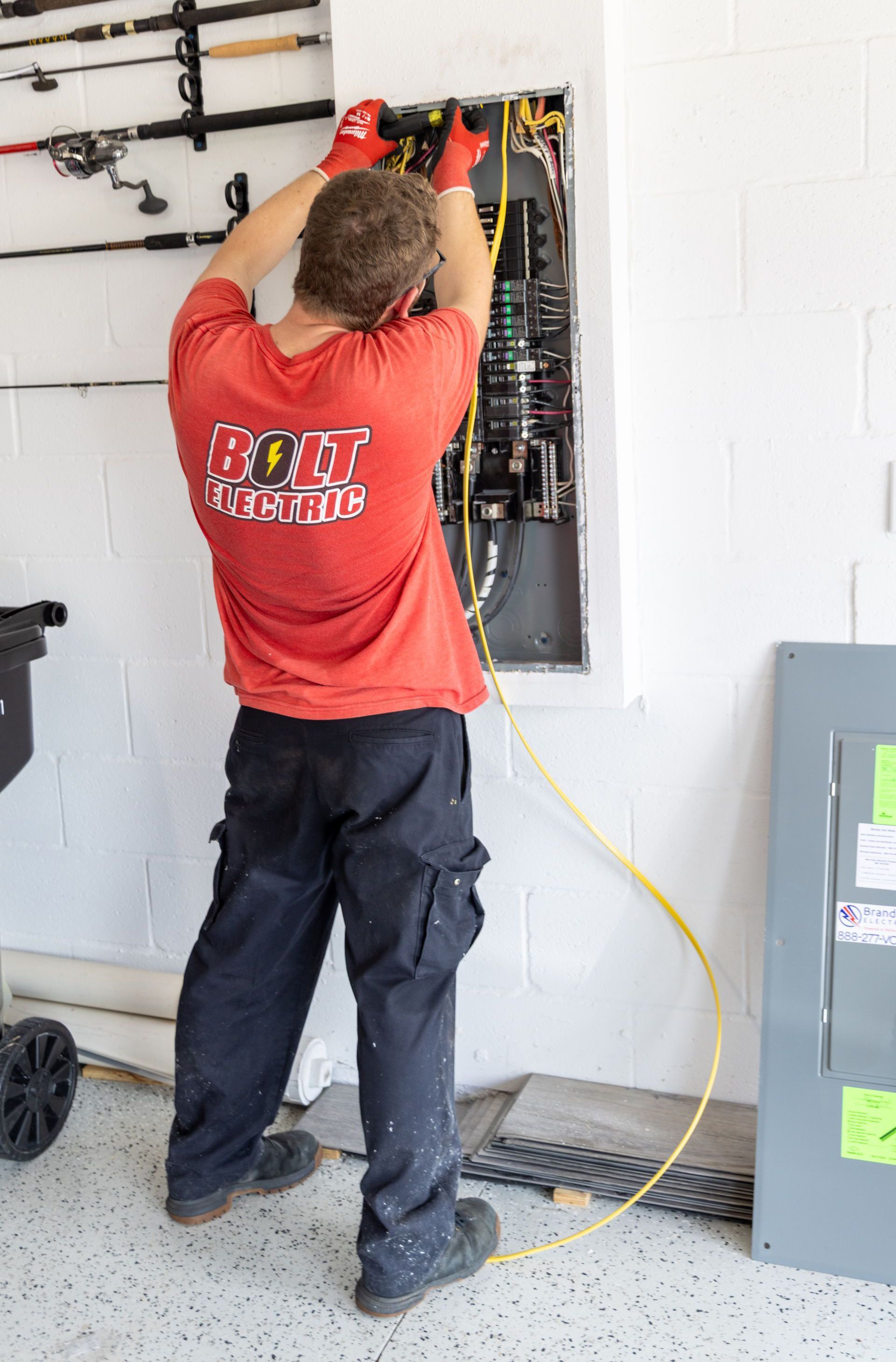 An electrician in a red shirt and work pants is working on an electrical panel on a white wall.