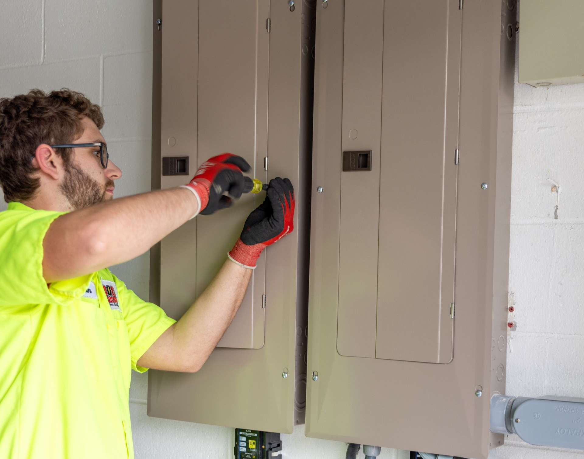An electrician, wearing safety gear, uses a screwdriver on an electrical panel.