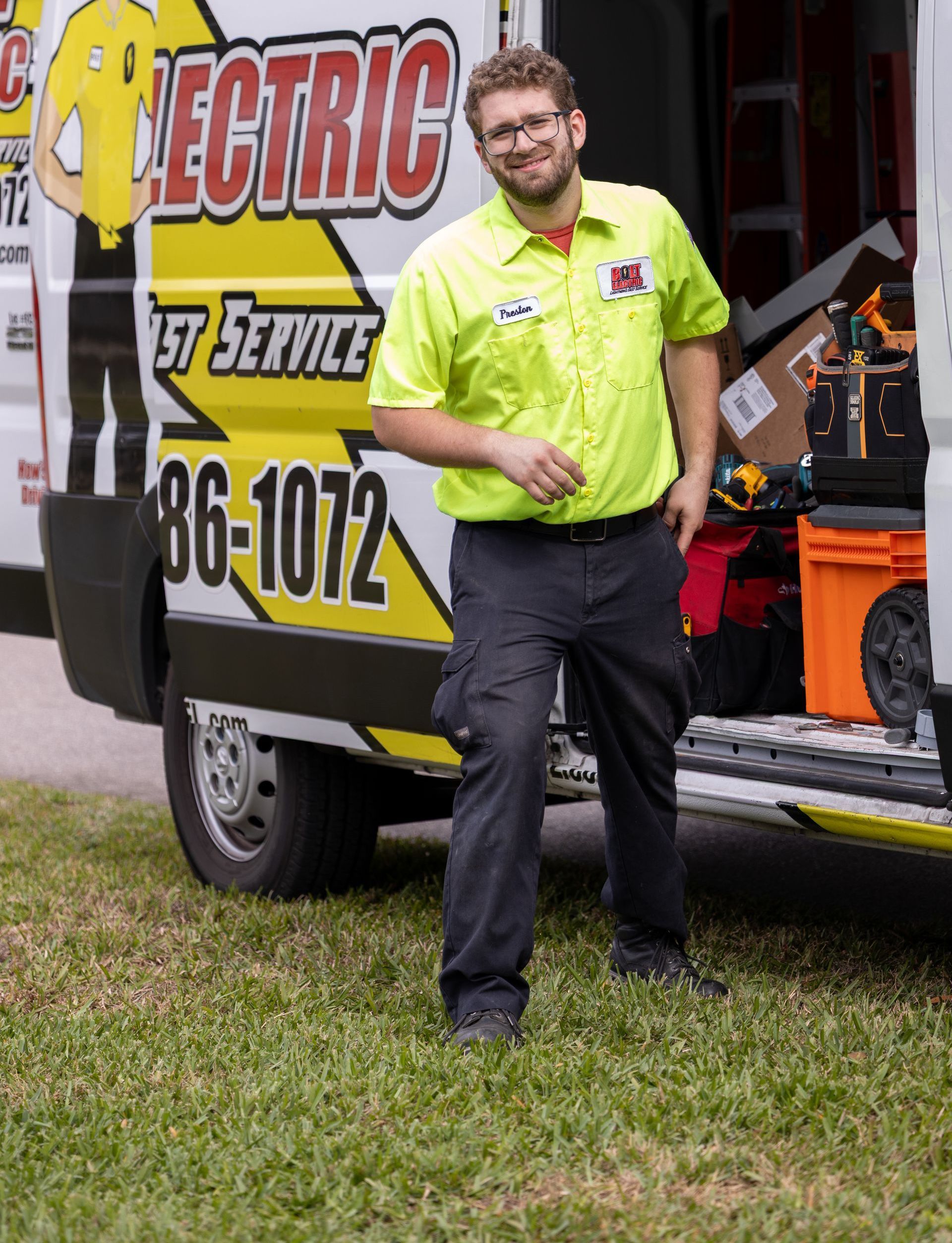 An electrician in a neon green shirt stands beside his company van.