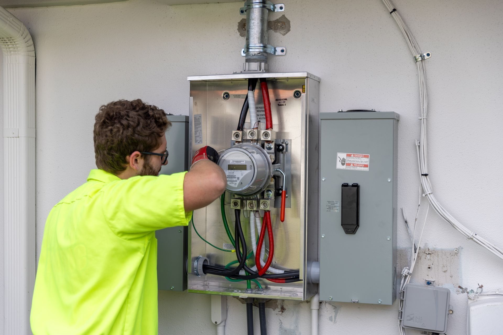 An electrician in a neon shirt, working on a gray electrical meter box mounted on a white wall.